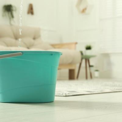 Close up of turquoise-colored plastic bucket on floor in living room with water dripping from ceiling into it.