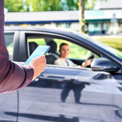 Arm of person in focus in the foreground, holding smartphone, with blurred dark car pulling up in the background, driver visible through the passenger window.