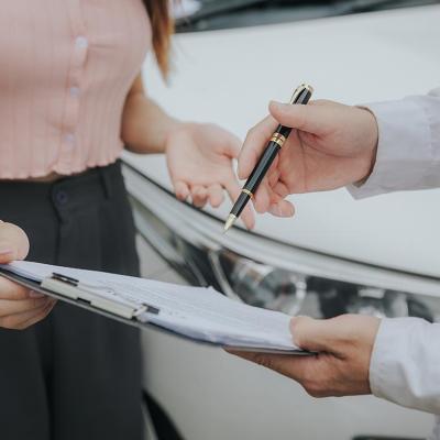 Close up of midsections of two people whose faces are not visible looking at a clipboard with forms on it with a white car in the background. 