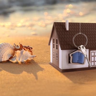 A model of a house with keys on a sandy beach beside seashells.
