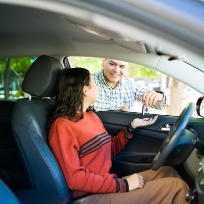 A teenage daughter inside a car receiving its keys from her proud dad.