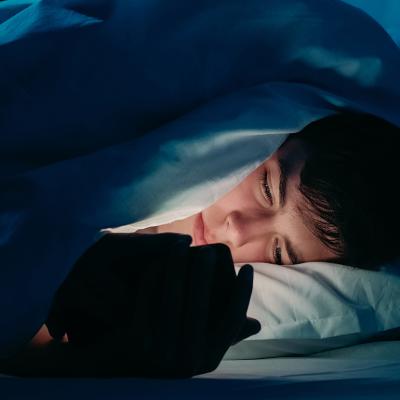 A male teenager using his smartphone under bedsheets at night.