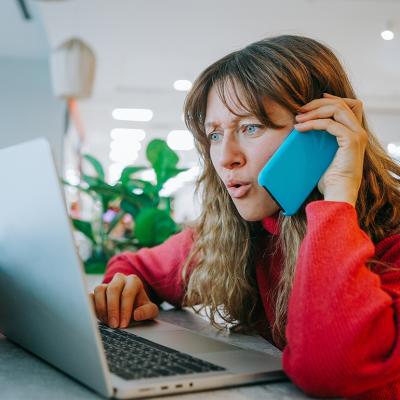 A woman talking on her smartphone while also booking flights using her laptop.