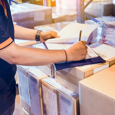 Warehouse worker writing on a clipboard on top of boxes as part of checking inventory.