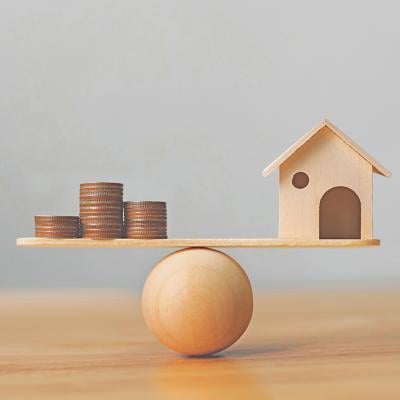 Wooden scale balancing a mini wooden figure of a house on the right and stack of coins on the left.
