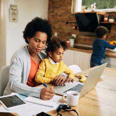 A black mother takes notes as she works at home while her young daughter sits on her lap.