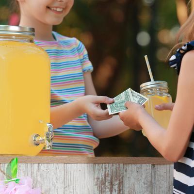 A young girl sells lemonade to another young girl at the park.