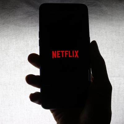 The Netflix logo on the screen of a mobile phone held by a person against a white background.