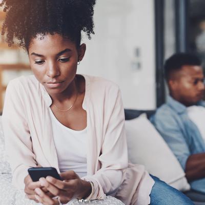 A black couple on their individual phones while sitting apart on the same couch at home.
