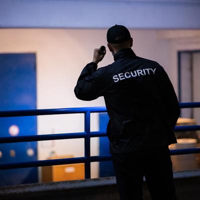 A person standing on a loading ramp outdoors at night wearing dark clothes with the word "SECURITY" displayed on the back of a shirt shines a flashlight toward a white wall with blue doors.