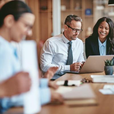 Two diverse businesspeople smiling while working on a laptop together at the end of a boardroom table in an office.
