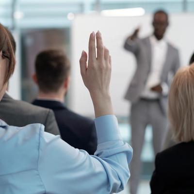 Rear view of raised hand with pointy nails of young employee wishing to answer question of lecturer while sitting behind group of other speakers at conference.
