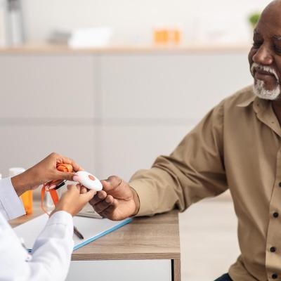 Portrait of black medical worker holding personal alarm button and giving it to mature patient, sitting at table in office, explaining how to use medical alert.