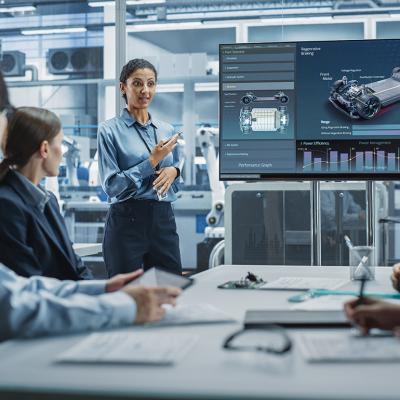 A female project manager leads her team of engineers with a presentation of a new car model in an EV manufacturing facility office.