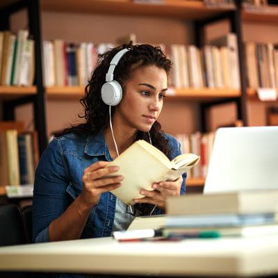 A fema collge student reading a book in a library while listening to music.