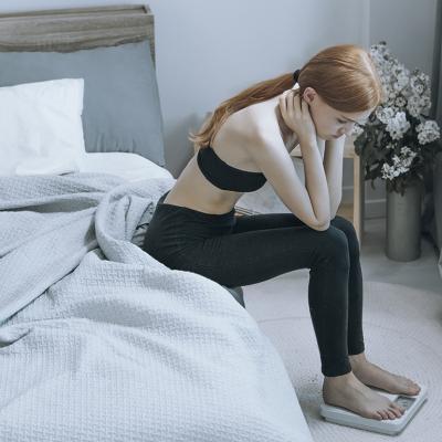A young woman sits by her bed while pensively checking weight on a scale.