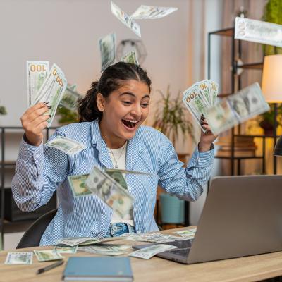 Young person throwing money and bills in the air in front of a computer; concept of getting rich quickly myth