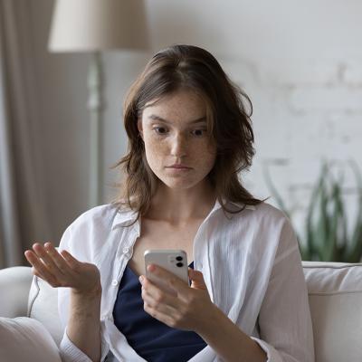 A woman sitting on a couch at home looking concerned at her phone.