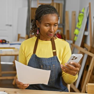 A black female carpentry business owner holds a document and reads information from her smartphone.