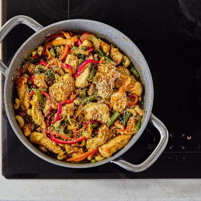 A colorful mix of stir-fried chicken, fresh vegetables, and cashews is sizzling in a pot on an induction cooktop. 
