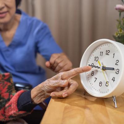 Close up of Asian elderly woman's hand pointing at analog clock with caregiver visible behind her.