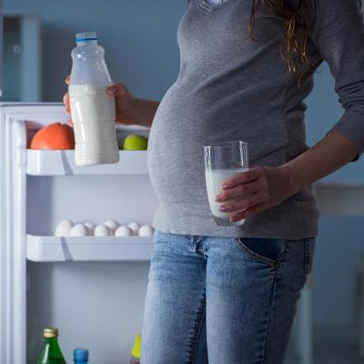 Pregnant woman, whose face is not shown, is illuminated by light of open fridge holding a glass in one hand and bottle of milk in the other.