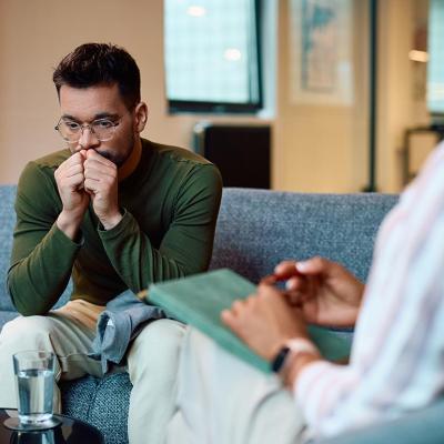Man in a long sleeve green shirt wearing glasses sits on a blue couch breathing into his hands. A box of tissues is on the table and a therapist's back is in the foreground.