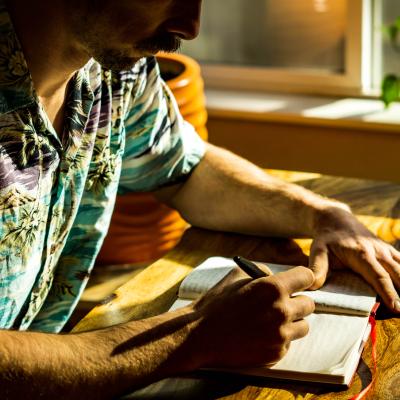 Man wearing a Hawaiian-style shirt sitting next to the window with light coming in, writing in a journal. 