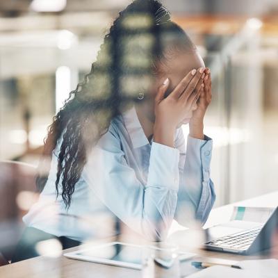 A stressed young black woman with her hands over her face at work.