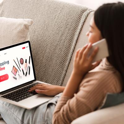 Back view of a woman sitting at home, on the phone, and browsing a shopping website on a laptop.