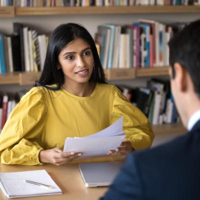 A woman behind a table holding papers and talking to a man sitting on the other side.