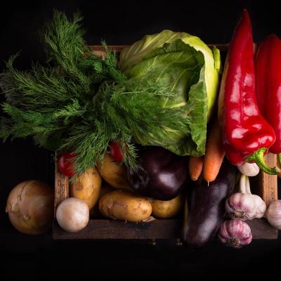 Assortment of beautifully-lit, fresh raw vegetables inlcuding leafy greens and red peppers in a wooden box against black background.