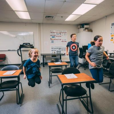 Three elementary-school-age students jumping in the air next to desks in a classroom. 