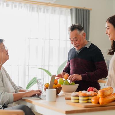 Mother father and adult daughter at the kitchen table talking with window in the background and fruit on the table.