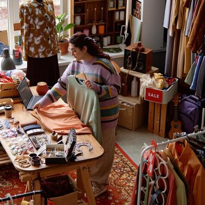 High-angle shot of sales assistant working at checkout desk cluttered with fashion accessories at second hand shop with retro style carpet selling clothes and household goods.