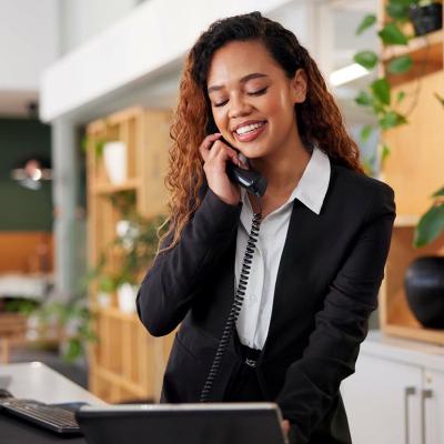 Customer service concierge answering phone standing behind reception desk looking at computer screen with modern decor and houseplants behind her.