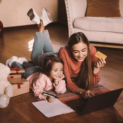 Girl writing wishlist, next to adult woman, both are laying on a rug looking a laptop online shopping while the woman is holding a credit card. There are wrapped gifts next to them along with a teddy bear.