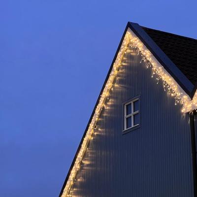 Icicle Christmas Lights hanging along the roof line of a wooden-fronted house. There is a small white window on the house, and a dusky blue sky behind the house.