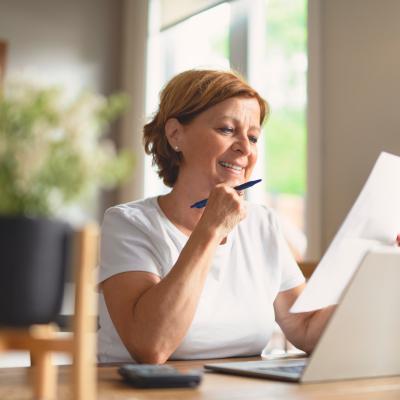 Middle-aged woman sitting at a table looking over paperwork with laptop open and pen in hand.