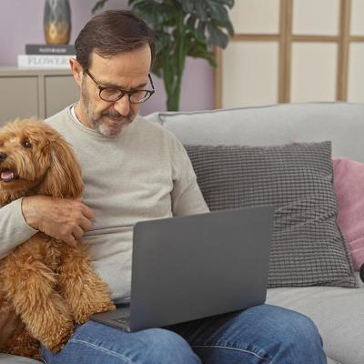 A hispanic middle-aged man cuddles a poodle while using a laptop in a cozy living room setting.