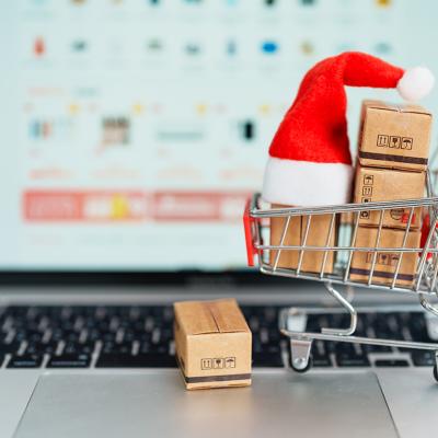 A small shopping cart figure with boxes and a Christmas hat, in front of a laptop displaying a shopping page.