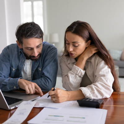 Serious young to middle aged couple discussing expenses at table with papers, a laptop and a calculator.