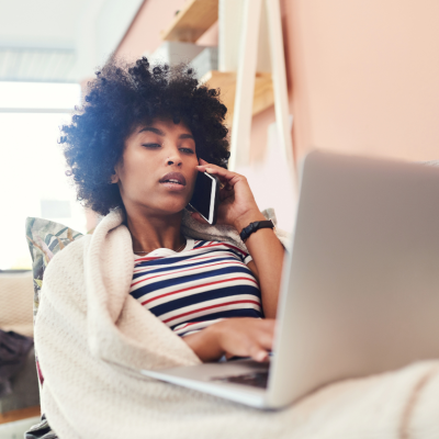A woman at home on the couch, wrapped in a blanket talking on the phone with a laptop on her lap.