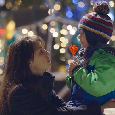 A mother kneels to the height of her child ourdoors and looks into his eyes, he is eating a heart-shaped lollipop and wearing a knitted cap with a pom pom in front of blurry christmas lights in the background.