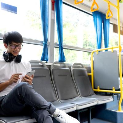 A college-age person wearing a crisp white shirt and headphones around his neck and bright white shoes sits on the side-facing seats in public transportation looking attentively at phone.