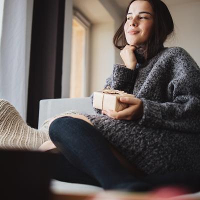 A young woman cozily sitting on a sofa at home holding a small gift box.