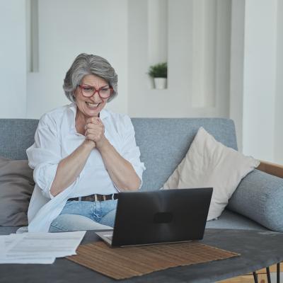 A senior woman sitting on a sofa and excitingly awaits good news on her laptop.