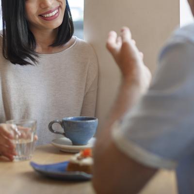 Two people sitting across from each other at a table in a cafe with water, a piece of cake, a mug in front of them. Just one person's smiling face is partially in view. 