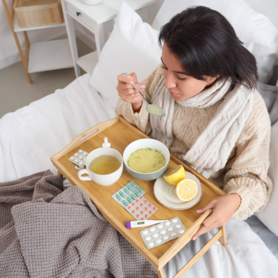 A woman sits in bed holding a spoon full of soup with a tray over her lap containing pills, nasal spray, a thermometer, lemons, a bowl of soup, and a mug of tea. 