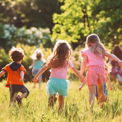 Young children running and playing in a sun-filled meadow with long grass and trees in the distance.